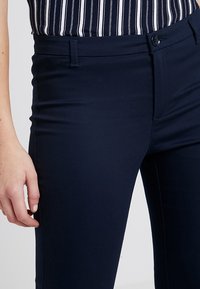 Mörkblå slim fit-byxor i stretchigt material, med knäppning, bältesöglor och en slät yta. Hand vilande på höften.