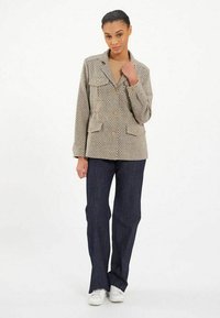 Veste à motif beige avec deux poches avant et des boutons en bois, associée à un jean évasé bleu foncé et des baskets blanches. Fond neutre.