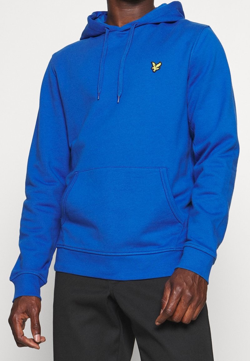 Uomo che indossa un felpa blu brillante con un logo di un uccello giallo sul petto e pantaloni neri, in piedi su uno sfondo semplice.
