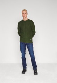 Dark green knitted sweater with a textured pattern, crew neckline, and ribbed cuffs, paired with blue jeans and black shoes.