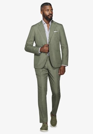 Mężczyzna w oliwkowozielonym garniturze, w paskowaną koszulę i pasujące mokasyny, idący, poprawiający guzik marynarki, na jednolitym białym tle.