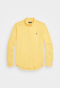 Camisa amarilla de manga larga, con botones y cuello, que presenta un pequeño logo oscuro en el lado izquierdo del pecho, dispuesta sobre un fondo blanco.