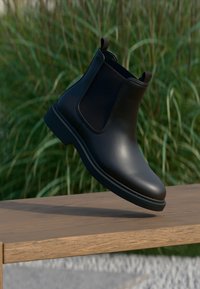 Black leather Chelsea boot with elastic side panels and pull tabs, suspended above a wooden surface with green foliage background.