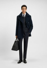 Cappotto di lana doppiopetto blu navale su un completo grigio, abbinato a scarpe in pelle nere e a una borsa nera testurizzata, in piedi di fronte a uno sfondo neutro.