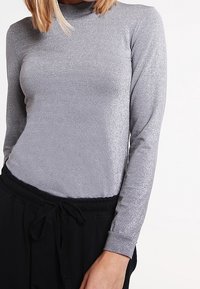 Harmaa pitkähihaine paita, joka on valmistettu kimaltavasta materiaalista ja jossa on korkea kaula-aukko, yhdistettynä löysiin mustiin housuihin, joissa on solmittava vyötärö.