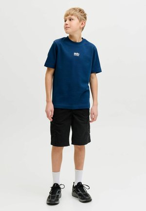 Junge trägt ein blaues kurzärmeliges T-Shirt, schwarze Cargoshorts, weiße Socken und schwarze Turnschuhe, steht und schaut zur Seite.