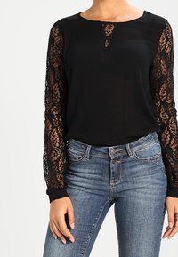 Schwarze Bluse mit langen Spitzenärmeln, rundem Ausschnitt und einem kleinen Ausschnitt an der Brust. Kombiniert mit blauen Jeans.