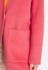 Rosa Wollmantel mit einer großen Vorderstasche, strukturiertem Schnitt, klaren Kanten und einer weichen Textur, präsentiert mit einem gemusterten Kleidungsstück darunter.