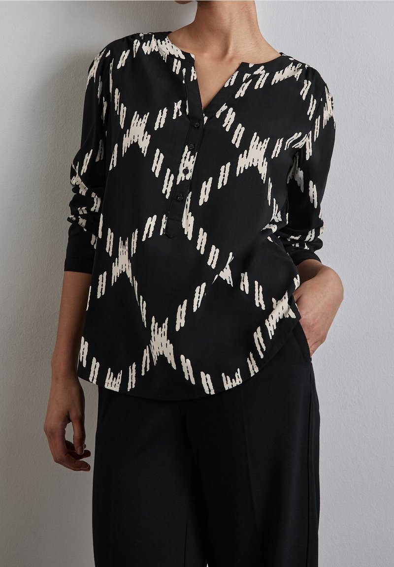 Blusa nera con motivo astratto bianco, caratterizzata da un collo a V con bottoni, maniche lunghe e tessuto liscio. Abbinata a pantaloni neri.