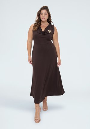 Vrouw draagt een mouwloze donkerbruine maxi-jurk met een geplooide halslijn en een broche, lopend naar voren in beige sandalen met hakken op een witte achtergrond.