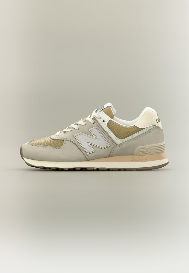 Μπέζ και καφέ New Balance αθλητικό παπούτσι με λευκά κορδόνια, διχτυωτές και σουέντ επιφάνειες και μαλακή σόλα σε ουδέτερο φόντο.