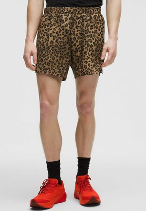 Mann trägt braune Shorts mit Leopardenmuster, schwarze Socken und leuchtend rote Turnschuhe, steht vor einem schlichten grauen Hintergrund.