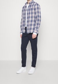 Ruudullinen vaaleanbeige paita, jossa on sinisiä ja mustia kuvioita, yhdistetty tummiin housuihin ja valkoisiin lenkkareihin, seisoo yksinkertaista taustaa vasten.