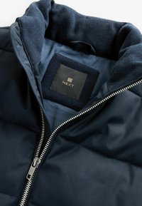 Navyblauwe gewatteerde jas met een rits sluiting, fleece-gevoerde kraag en een vierkant label met "NEXT" in gouden tekst. Glad oppervlak.