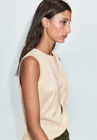 Top senza maniche beige in tessuto morbido con scollatura a girocollo, abbinato a una lunga collana dorata. Design semplice, con motivi minimalisti.