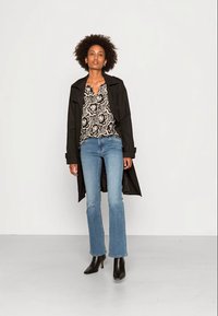 Casaco longo preto sobre uma blusa estampada, jeans azuis claros e evasê, e botins pretos de salto alto. Texturas suaves e detalhes de design floral.