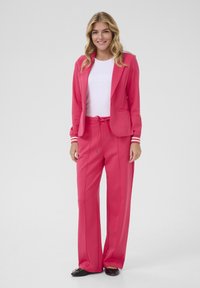Un costume rose composé d'un blazer ajusté avec des accents à rayures blanches et un pantalon large avec une taille à cordon, assorti d'un haut blanc.