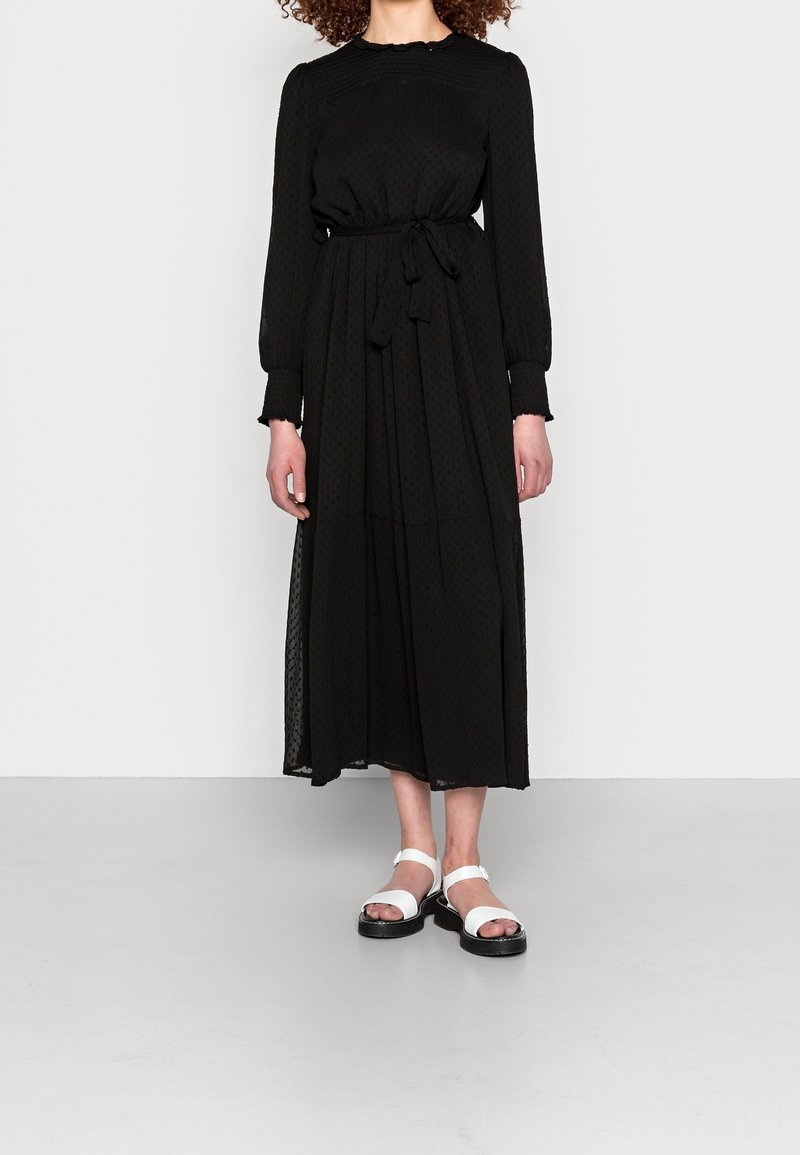Femme portant une robe midi noire à manches longues avec une ceinture nouée à la taille et des sandales blanches à bouts ouverts, debout sur un fond uni.