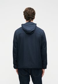 Άνδρας με κοντά σγουρά μαλλιά, φορώντας ναυτικό μπουφάν με κουκούλα και σκούρα τζιν, στέκεται με το βλέμμα στραμμένο σε έναν απλό λευκό φόντο.