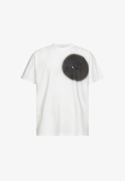 T-shirt blanc à manches courtes avec un cercle noir au pochoir sur le côté gauche de la poitrine et une petite poche partiellement visible à l'intérieur du cercle.
