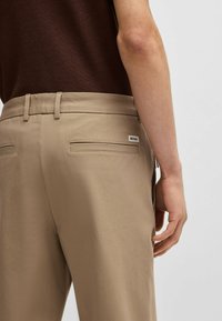 Beżowe spodnie o gładkiej fakturze, z dwoma tylnymi kieszeniami i widoczną metką marki. Pas jest płaski z szlufkami na pasek.