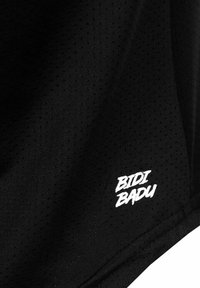 Tela negra transpirable con pequeñas perforaciones y logo "BIDI BADU" impreso cerca del dobladillo inferior.
