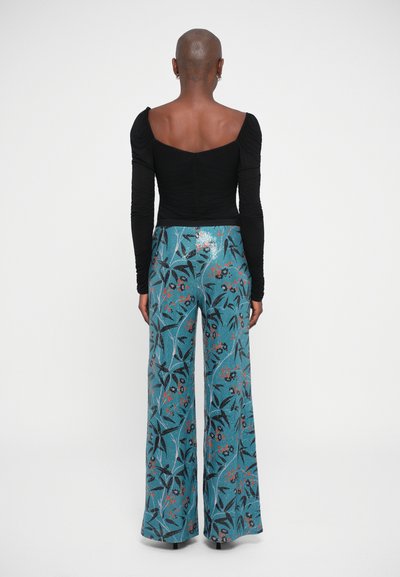 Μαύρη μπλούζα με μακριά μανίκια σε συνδυασμό με τυρκουάζ παντελόνι σε φαρδιά γραμμή που έχει λουλουδάτα μοτίβα και γυαλιστερά αξεσουάρ, εμφανιζόμενο από πίσω.