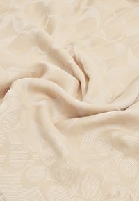 Tissu beige tissé avec un motif texturé de formes qui se chevauchent. Doux, se drape fluidement, présentant des variations tonales subtiles et des bords à franges.