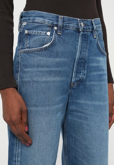 Jeans de tiro alto en un lavado medio de color azul. Incluyen cinco bolsillos, cierre frontal con botón y un ligero desgaste. Tejido de denim texturizado.