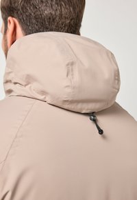 Uomo che indossa un giacca beige con cappuccio regolabile, visto di schiena e leggermente di lato su uno sfondo semplice.