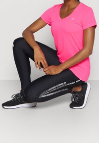 Růžové sportovní tričko s výstřihem do V v kombinaci s černými legínami s bílým logem "Under Armour". Model má na sobě černé sportovní boty.