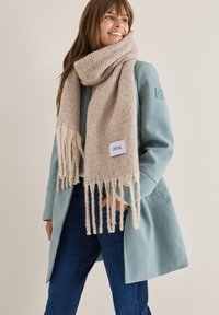 Beige stickad scarf med fransdetaljer hängande runt halsen, kombinerad med en ljusblå rock. Scarfen har en liten etikett som står "CECIL."
