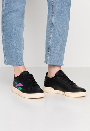 Person wearing light blue jeans and black sneakers with purple and green accents, standing on a plain gray floor.