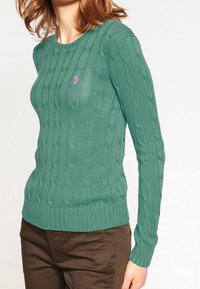 Grön stickad tröja med ett kabelmönster, rund halsringning och långa ärmar. Har en liten rosa broderad logotyp på bröstet.