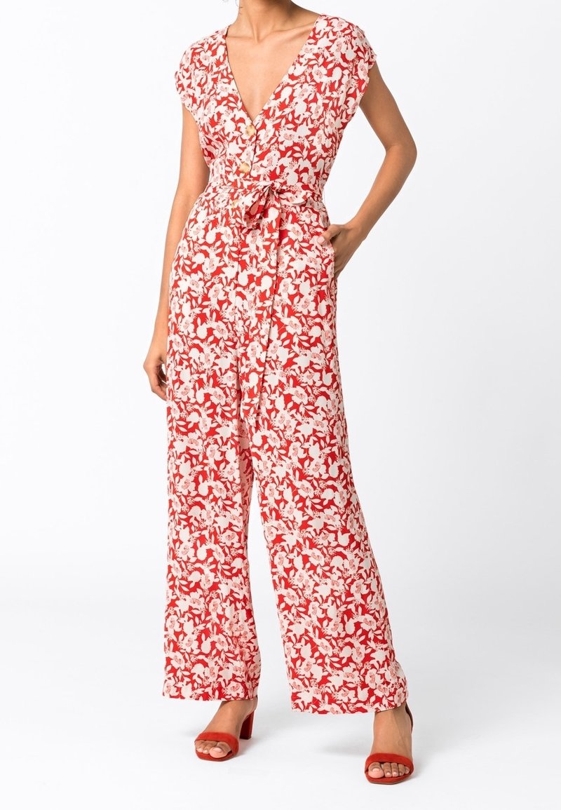 Femme portant une combinaison pantalon large à fleurs rouges et blanches avec un col en V, des boutons, une ceinture à nouer et des talons rouges ouverts à l'avant.