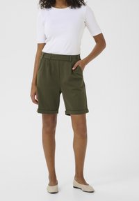 Shorts vert olive avec ourlets retournés, coupe droite et plis. Associés à une chemise blanche ajustée et des ballerines beige.
