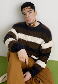 Strickpullover in dunkelbraun, creme und marineblauen Streifen mit einer strukturierten Oberfläche. Das Modell trägt lockere, helle Hosen und sitzt vor einem grünen Hintergrund.