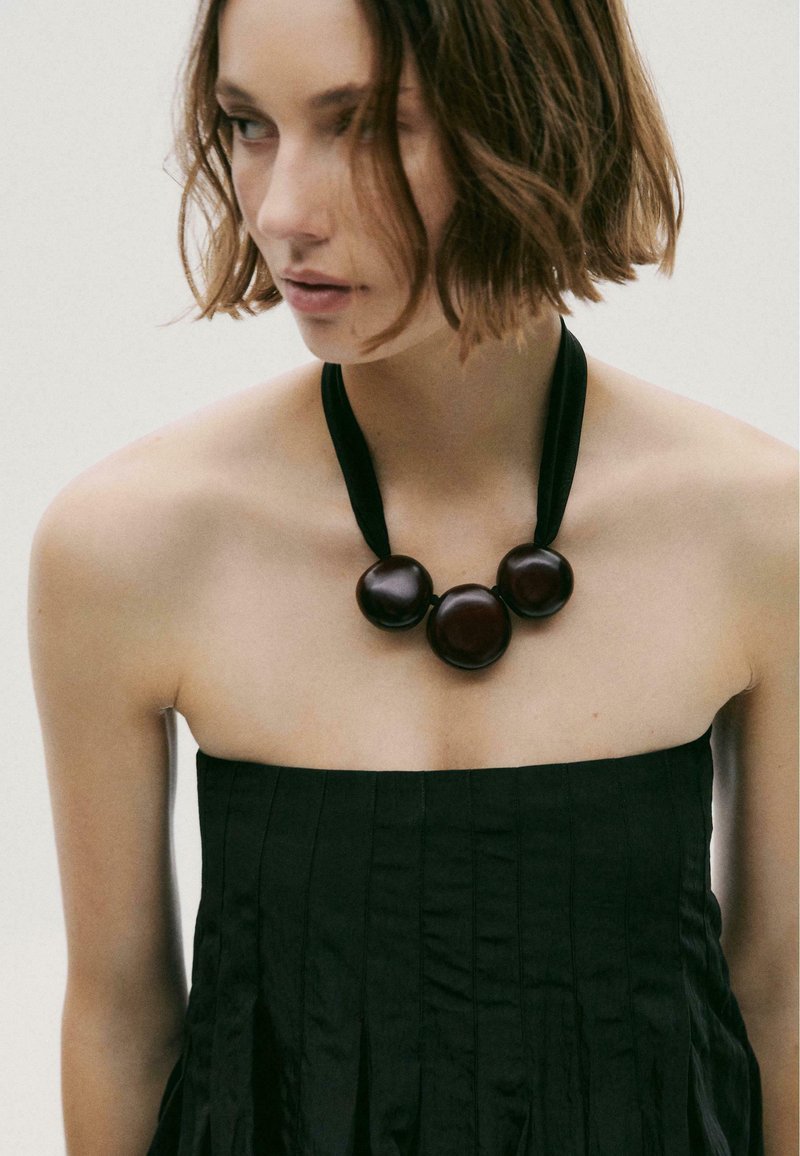 Collana in tessuto nero con tre grandi palline di legno scuro. Texture liscia e finitura opaca, indossata sopra un top senza spalline a pieghe.