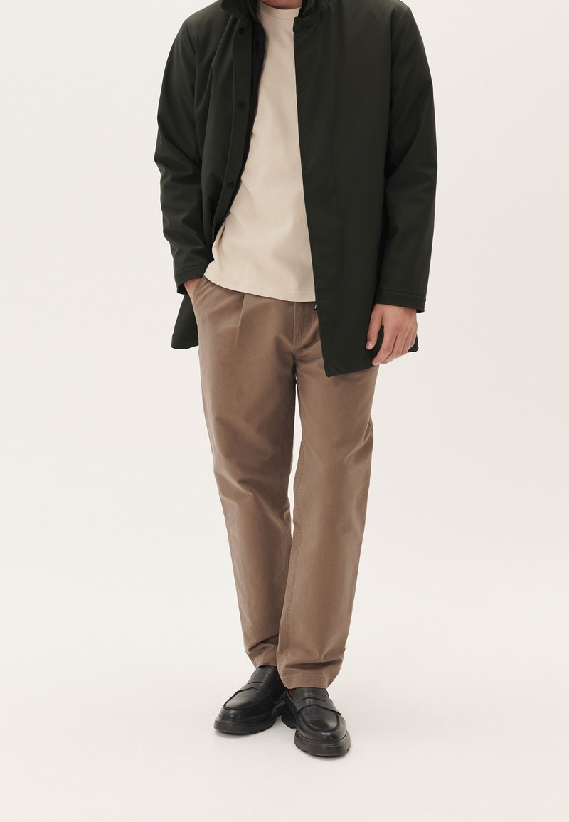 Homme portant un manteau noir, une chemise beige, un pantalon marron et des chaussures noires, debout avec une main dans la poche sur un fond uni.