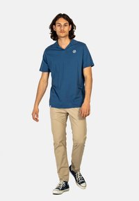 Camisa polo de manga corta azul con cuello, que presenta un pequeño logo en el pecho, combinada con pantalones beiges y zapatillas azules.