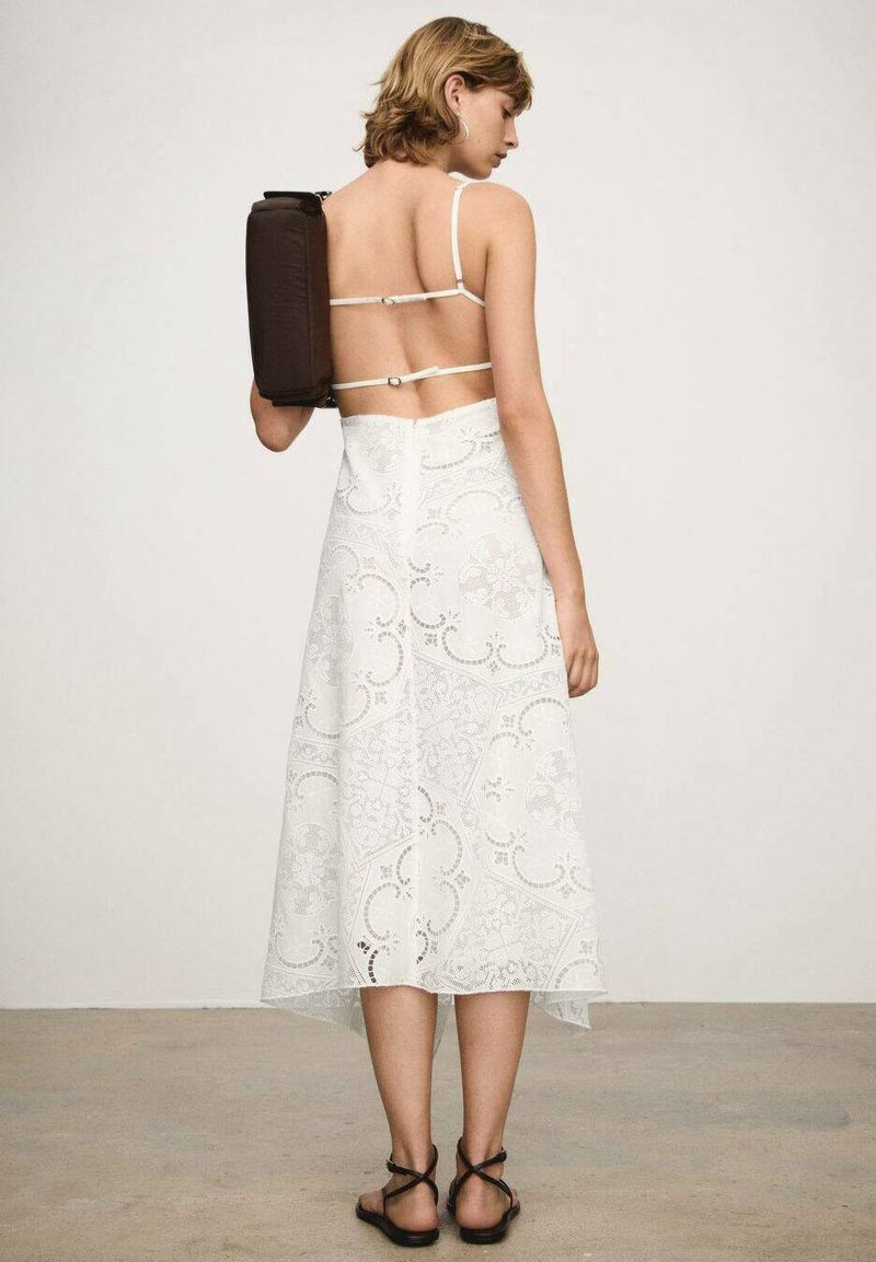 Biała koronkowa sukienka z przezroczystym tyłem, regulowanymi ramiączkami na sznurki i asymetrycznym dołem. Ciemnobrązowa torebka noszona na jednym ramieniu. Widoczne czarne sandały.