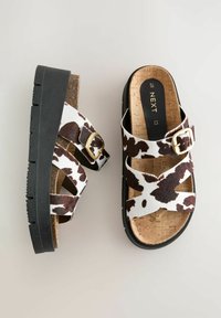 Sandalias de estampado de vaca en marrón y blanco con una plantilla de corcho y suela de plataforma negra. Presentan correas ajustables y un detalle de hebilla dorada.