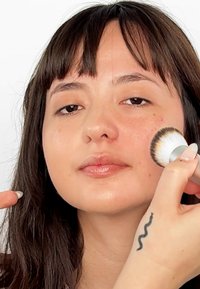 En sminkebørste med et sølvhåndtak brukes på et ansikt som viser hudfeil; modellen har langt mørkt hår og en tatovering på håndleddet.