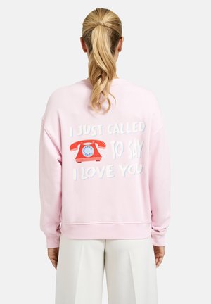 Blonde Frau mit Pferdeschwanz, die ein hellrosa Sweatshirt trägt, auf dessen Rückseite ein rotes Wählscheibentelefon und der Text „ICH HABE NUR ANGERUFEN, UM DIR ZU SAGEN, DASS ICH DICH LIEBE“ abgebildet sind.