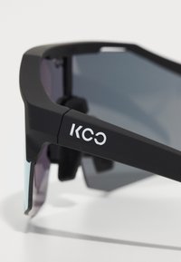 Black sunglasses with a matte frame, tinted lenses, and "KCC" logo on the side. Features a curved design and sleek texture.