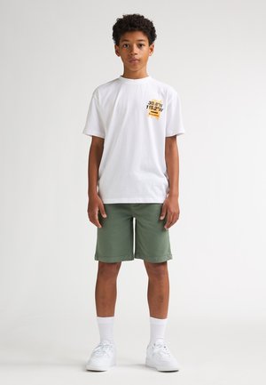 T-shirt bianca in cotone con stampa grafica nera e gialla; abbinata a pantaloncini verdi in cotone, sneaker bianche e calzini bianchi.