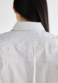 Camicetta a righe bianche e navy con colletto, caratterizzata da lettere ricamate "DKNY" in bianco sul retro, che mostra una texture liscia.