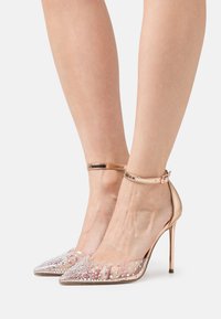 Scarpe con tacco alto trasparenti, caratterizzate da una tomaia in plastica trasparente adornata con borchie argentate, punta affusolata e una cinghia alla caviglia in metallizzato oro rosa.