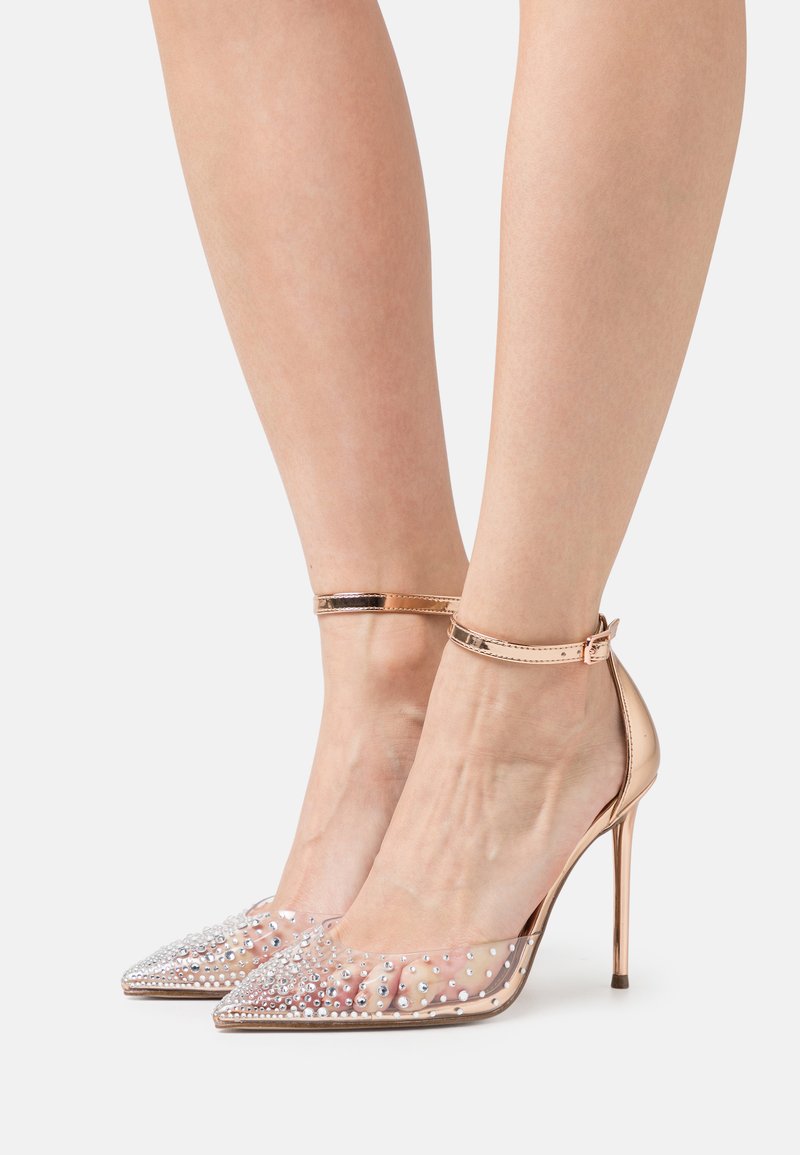 Scarpe con tacco alto trasparenti, caratterizzate da una tomaia in plastica trasparente adornata con borchie argentate, punta affusolata e una cinghia alla caviglia in metallizzato oro rosa.
