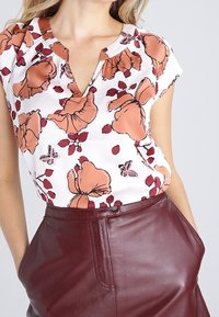 Blouse blanche à manches courtes ornée d'un motif floral en orange et bordeaux, avec un décolleté en V et une coupe décontractée. Associée à une jupe en cuir bordeaux.